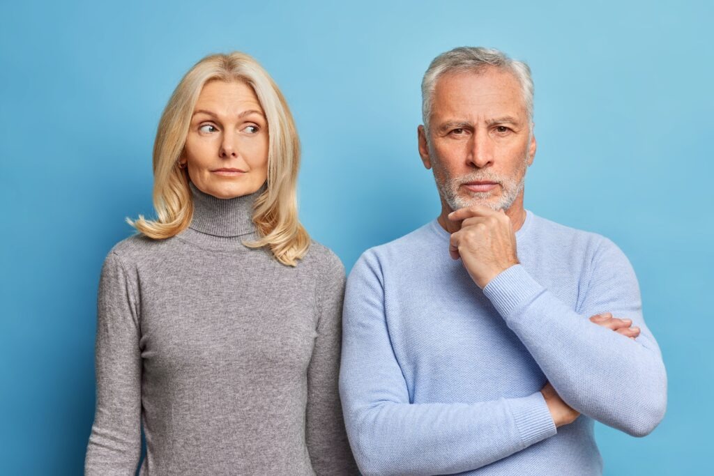 Serious mature woman and man stand closely to each other have thoughtful expressions dressed in casual clothes isolated over blue background. Self confident bearded male pensioner holds chin