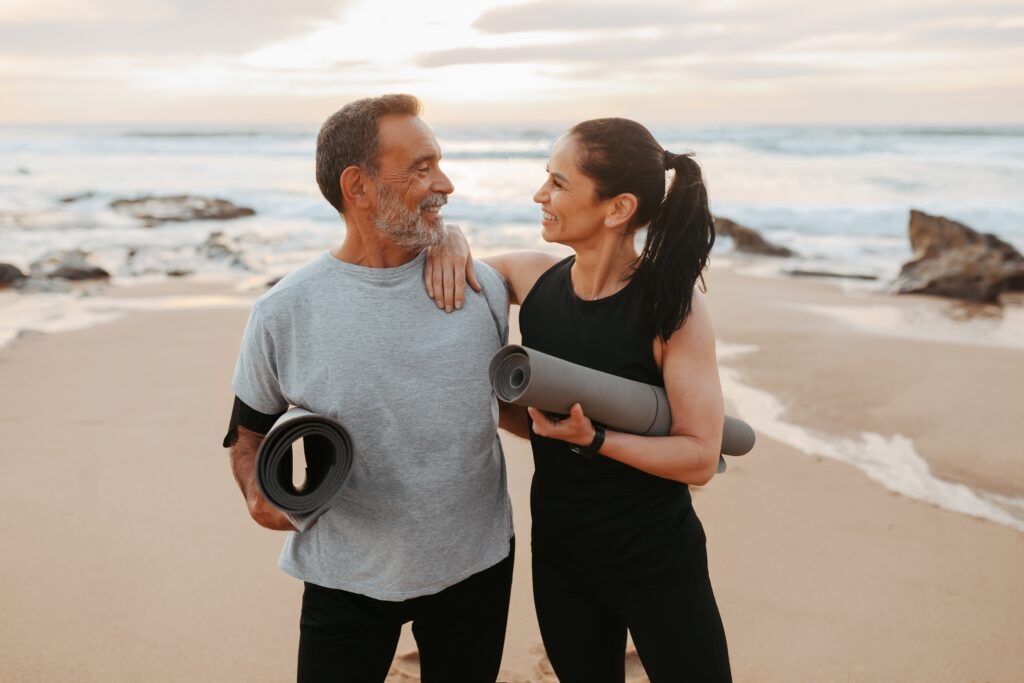 Smiling mature european couple in sportswear with mats enjoy workout together in morning on sea beach, outdoor. Active lifestyle at spare time, sports, fitness and body care