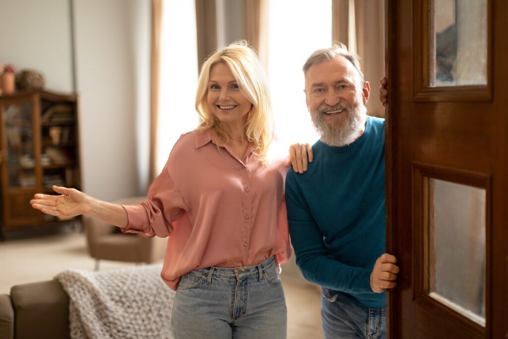 Cheerful Senior Spouses Opening Door Of New Apartment, Welcoming You Smiling To Camera Hugging At Home. Couple Standing In Doors Of Their House. Real Estate Offer And Ownership Concept