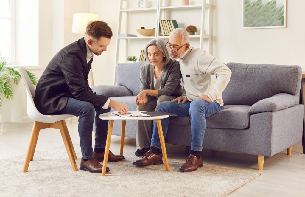 Elderly couple consults with advisor about investments or real estate planning in comfort of home. Young male professional is discussing documents with attentive mature couple in cozy living room.