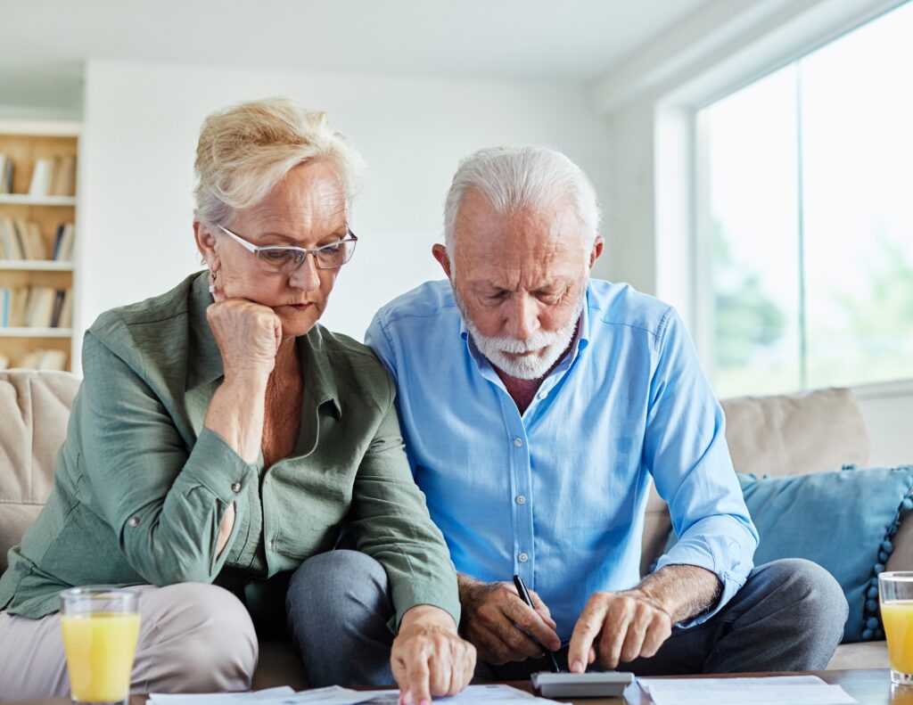 Senior couple checking bills and calculating expenses using a calculator at home, debt taxes and home budget concept, worried married elderly couple